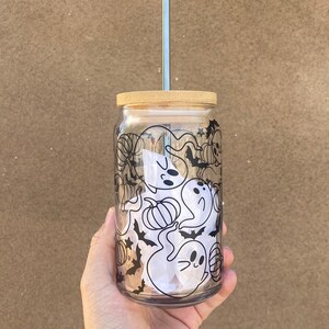 以下が含まれることがあります： 竹製の蓋と銀色のストローが付いた透明なガラスタンブラー。タンブラーは、お化け、カボチャ、コウモリを特徴とする黒いハロウィーンをテーマにしたデザインで装飾されています。