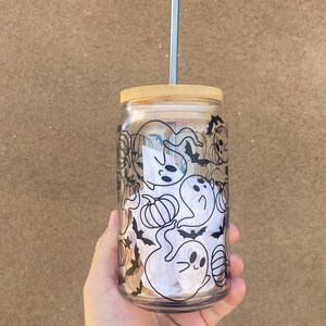 以下が含まれることがあります： 竹製の蓋と銀色のストローが付いた透明なガラスタンブラー。タンブラーは、お化け、カボチャ、コウモリを特徴とする白黒のハロウィーンデザインで装飾されています。タンブラーは手で持たれています。