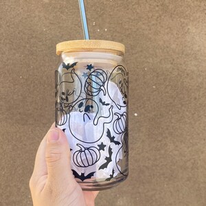 以下が含まれることがあります： 竹製の蓋と銀色のストローが付いた透明なガラスタンブラー。ガラスには、幽霊、カボチャ、コウモリ、星など、ハロウィーンをテーマにした黒いグラフィックが施されています。タンブラーは手で持たれています。