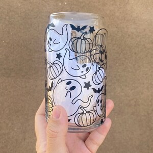 以下が含まれることがあります： ハロウィーンをテーマにしたデザインの、透明なガラス製の缶型グラス。デザインは、幽霊、カボチャ、コウモリ、星の黒いイラストが特徴です。グラスは手で持たれています。