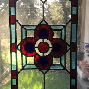 Puede incluir: Un panel de vitral con un diseño geométrico que presenta círculos rojos, azules y blancos en el centro. El panel está enmarcado con un borde negro y tiene un borde azul claro y rojo.