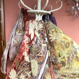 Pu&ograve; includere: Quattro sciarpe con stampa botanica sono appese a un appendiabiti bianco. Le sciarpe sono in tonalit&agrave; di marrone, giallo, arancione e rosso. Le sciarpe sono realizzate in un tessuto morbido e fluido.