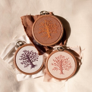 May include: Three embroidered hoop art pieces featuring trees in different colors. The hoops are made of wood and have a brown finish. The trees are embroidered in orange, purple, and pink thread.