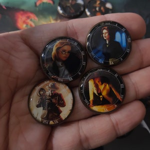 May include: Four round, gold-rimmed, clock-face style tokens with different images of people. The tokens are on a hand.