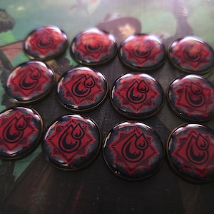 May include: Twelve circular tokens with a glossy finish. Each token displays a red emblem with a teardrop design on a dark grey background, framed by a gold-coloured metal rim. The tokens are arranged on a patterned surface.
