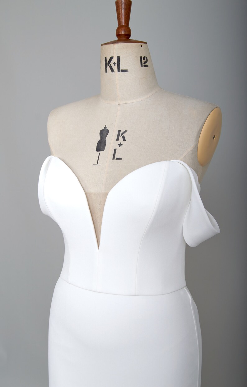 K&ouml;nnte beinhalten: Ein wei&szlig;es, schulterfreies Kleid mit einem herzf&ouml;rmigen Ausschnitt und einer figurbetonten Silhouette. Das Kleid wird auf einer Schaufensterpuppe pr&auml;sentiert.