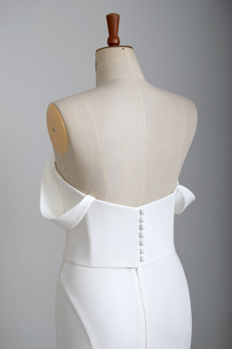 K&ouml;nnte beinhalten: Wei&szlig;es, schulterfreies Brautkleid mit einem figurbetonten Oberteil und einem langen Rock. Das Kleid hat eine Reihe von Kn&ouml;pfen auf der R&uuml;ckseite.