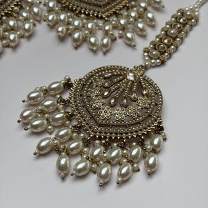 May include: An ornate gold-toned jewelry set featuring a central pendant and earrings, all adorned with faux pearls. The pendant and earrings have intricate designs with small, round pearls and larger, teardrop-shaped pearls hanging from the bottom.