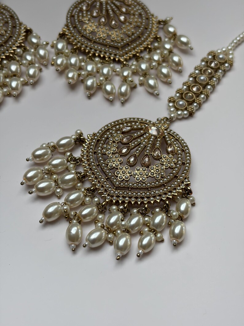 May include: An ornate gold-toned jewelry set featuring a central pendant and earrings, all adorned with faux pearls. The pendant and earrings have intricate designs with small, round pearls and larger, teardrop-shaped pearls hanging from the bottom.