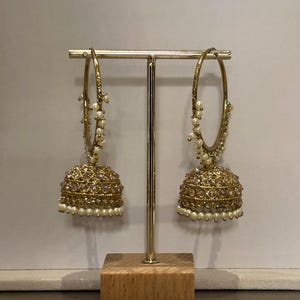 May include: Gold-toned hoop earrings with a bell-shaped drop. The earrings feature a detailed design with clear and pearl-like accents. They are displayed on a gold stand with a wooden base, against a neutral background.
