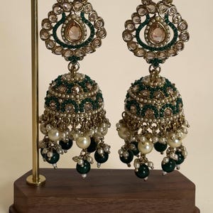 May include: A pair of gold-toned jhumka earrings featuring intricate green and clear stone embellishments, small white pearls, and dangling dark green beads. The earrings are suspended from a gold metal stand with a dark wooden base.