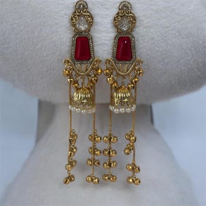 May include: A pair of gold-toned earrings with a rectangular red stone and a dangling design. The earrings feature a floral filigree design, small gold bells, and strands of gold beads and faux pearls.