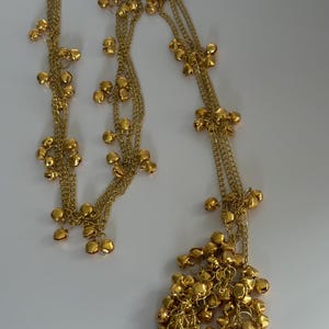 May include: A long gold-toned chain necklace featuring multiple strands adorned with numerous small, round metallic jingle bells. The bells are distributed along the length of the chains, with a dense cluster of bells gathered at the bottom end.