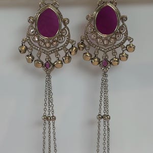 May include: A pair of ornate silver-toned earrings with a central oval of deep magenta. The earrings feature a detailed filigree design, small clear stones, and dangling chains with bell-shaped charms. The earrings are designed for pierced ears.