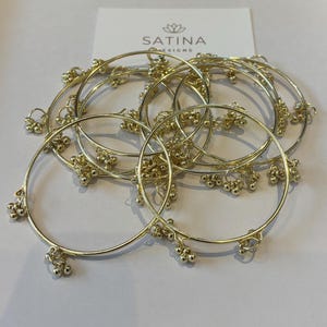 May include: Gold bangles with small dangling bead clusters. The bangles are arranged in a circular pattern, with the brand name "SATINA DESIGNS" printed on a white card in the background. The jewellery is likely for women.