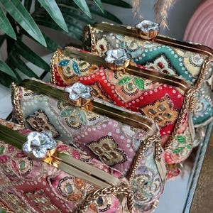 May include: A collection of four rectangular clutch bags featuring intricate embroidery, sequins, and small mirrors. Each bag has a gold-tone metal frame, a detachable chain strap, and a flower-shaped crystal clasp closure.