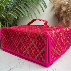 May include: A rectangular storage case with a top handle, featuring a vibrant magenta fabric exterior with a quilted diamond pattern. The surface is decorated with a repeating gold metallic floral motif and small orange dots within each diamond.