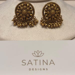 May include: A pair of antique gold-colored earrings. Each earring features a circular design with intricate floral patterns and small dangling beads. The earrings are displayed on a neutral-colored surface.