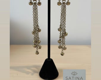 May include: Gold-toned dangle earrings with a floral design and dangling chains with small bell-shaped charms. The earrings are displayed on a black stand. The Satina Designs logo is visible.