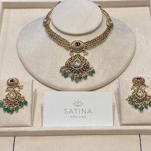 May include: A gold-coloured necklace and earring set. The necklace has a central pendant with a ruby and a teardrop-shaped white stone, with green beads. The earrings match the pendant design. The brand name "SATINA DESIGNS" is visible on a card.