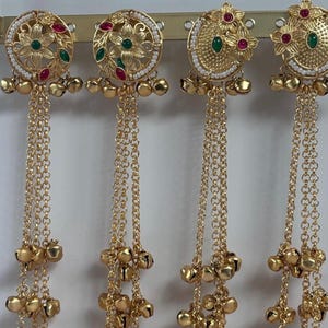 May include: Four gold-colored earrings with intricate floral designs and dangling chains. Each earring features a circular top adorned with red and green gemstones, surrounded by small white beads. The chains end with clusters of small bells.
