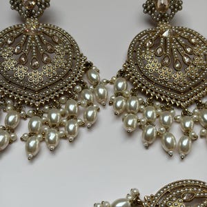 May include: Gold-toned, ornate earrings with a circular design, featuring faux pearls and clear crystals. The earrings have a floral-inspired top and dangling pearl clusters, creating a glamorous look. The jewelry is set against a white background.
