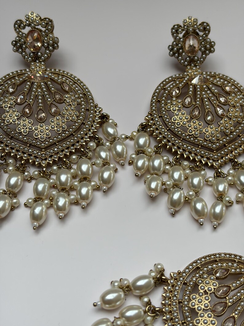 May include: Gold-toned, ornate earrings with a circular design, featuring faux pearls and clear crystals. The earrings have a floral-inspired top and dangling pearl clusters, creating a glamorous look. The jewelry is set against a white background.