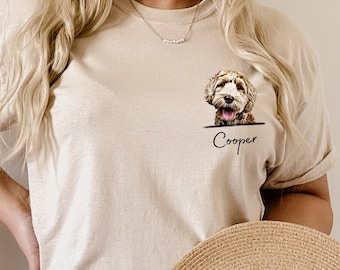 May include: Beige t-shirt featuring a dog portrait with the name "Cooper" printed below. The dog has brown and white fur. The person is holding a straw hat.