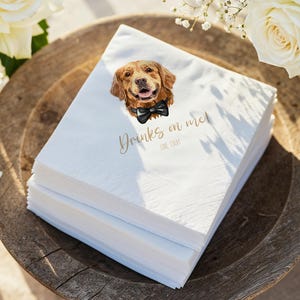 May include: A stack of white paper napkins with a watercolor illustration of a golden retriever wearing a black bow tie. The napkins have the text "Drinks on me!" in gold script. The napkins are on a wooden tray.