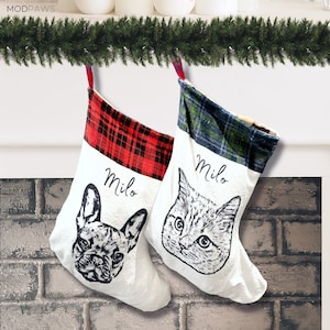以下が含まれることがあります： ペットの肖像画が描かれた白いクリスマスストッキング2つ。1つはフレンチブルドッグ、もう1つは猫です。どちらも「Milo」という名前と赤と緑のチェック柄のトップスが付いています。緑のガーランドが上に吊り下げられています。