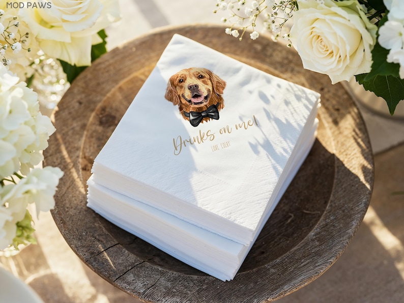 May include: A stack of white paper napkins with a watercolor illustration of a golden retriever wearing a black bow tie. The napkins have the text "Drinks on me!" in gold script. The napkins are on a wooden tray.