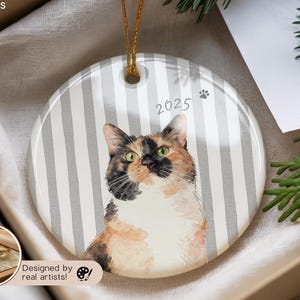 Pode incluir: Enfeite redondo de cer&acirc;mica com um retrato em aquarela de um gato calic&oacute; com olhos verdes. O enfeite tem um fundo listrado cinza e branco, o ano "2025" e uma pegada. Uma corda dourada &eacute; anexada para pendurar.