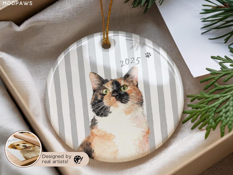 Pode incluir: Enfeite redondo de cer&acirc;mica com um retrato em aquarela de um gato calic&oacute; com olhos verdes. O enfeite tem um fundo listrado cinza e branco, o ano "2025" e uma pegada. Uma corda dourada &eacute; anexada para pendurar.
