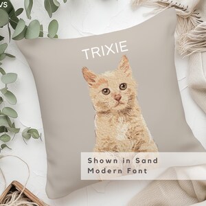 May include: A beige pillow with a light orange cat illustration and the name "TRIXIE" in a white sans-serif font. The text "Shown in Sand Modern Font" is also printed on the pillow.