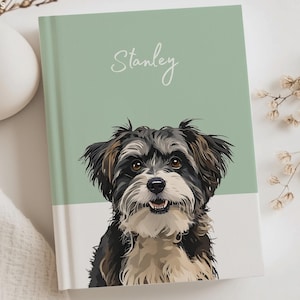 Puede incluir: Un cuaderno de tapa dura con una cubierta dividida en verde salvia y blanco, con una ilustración digital de un perro de pelo alborotado en negro, blanco y marrón. El nombre Stanley aparece en cursiva blanca sobre el retrato en la zona verde.
