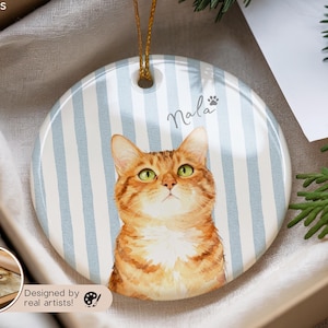 Pode incluir: Enfeite redondo de cer&acirc;mica com uma pintura em aquarela de um gato malhado laranja com olhos verdes. O enfeite tem um fundo listrado azul e branco e o nome "Nala" est&aacute; escrito acima do gato.