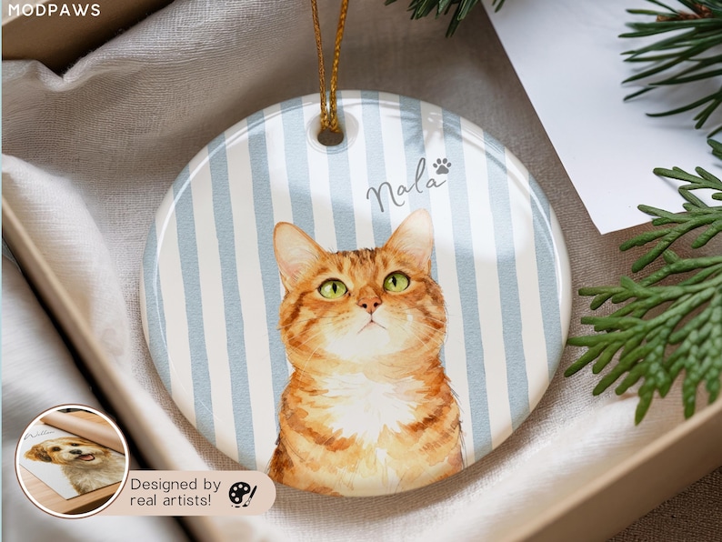 Pode incluir: Enfeite redondo de cer&acirc;mica com uma pintura em aquarela de um gato malhado laranja com olhos verdes. O enfeite tem um fundo listrado azul e branco e o nome "Nala" est&aacute; escrito acima do gato.