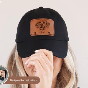 May include: Black baseball cap with a brown leather patch featuring a dog portrait, paw prints, and the name "Charlie." The cap is a classic style, suitable for casual wear. The phrase "Designed by real artists!" is visible.