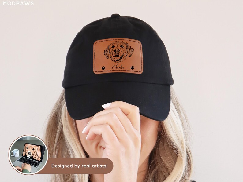 May include: Black baseball cap with a brown leather patch featuring a dog portrait, paw prints, and the name "Charlie." The cap is a classic style, suitable for casual wear. The phrase "Designed by real artists!" is visible.