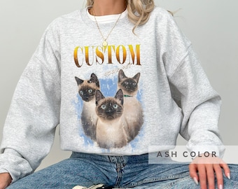 May include: An ash gray crewneck sweatshirt featuring a graphic print of three Siamese cats against a blue watercolor-style background. The text CUSTOM YOUR PET is printed in a bold, orange-to-yellow gradient font above the cat illustration.