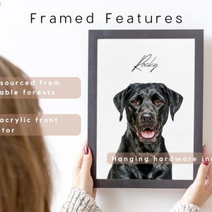 May include: A framed portrait of a black dog named Rocky, with a black frame. The image is held by a person. The frame features wood sourced from renewable forests, a clear acrylic front protector, and includes hanging hardware.