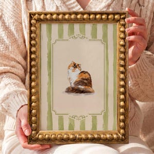 Puede incluir: Una lámina rectangular en un marco dorado con cuentas que presenta una ilustración de un gato calicó centrada sobre un fondo crema con rayas verticales verde salvia y bordes decorativos con volutas. El texto MOD PAWS aparece en la esquina superior izquierda.