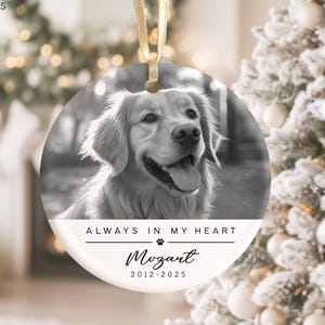 May include: A round, black and white pet memorial ornament with a photo of a Golden Retriever. The ornament has the text "ALWAYS IN MY HEART" and the name "Mozart" with the years 2012-2025. A gold ribbon is attached for hanging.