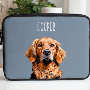 以下が含まれることがあります： ゴールデンレトリバー犬の肖像画が描かれたグレーのラップトップスリーブ。「COOPER」という犬の名前が肖像画の上に白い文字で書かれています。スリーブには黒いジッパーとトリムが付いています。