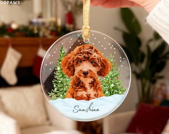 May include: A round, clear acrylic Christmas ornament featuring a printed illustration of a brown curly-haired dog sitting between two green pine trees. The design includes white snow, the text Sunshine, and a gold ribbon for hanging. MODPAWS is visible.