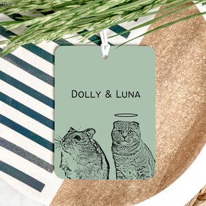 May include: A light green car air freshener with a black and white illustration of a cat and a hamster. The text "DOLLY & LUNA" is printed on the air freshener.