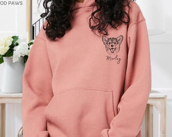 Puede incluir: Una sudadera con capucha de color rosa empolvado que presenta una ilustración de línea negra de la cara de un perro en el pecho izquierdo. Debajo de la ilustración del perro, el nombre Marley está impreso en cursiva negra. Incluye un bolsillo frontal.
