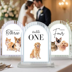 May include: Clear acrylic table number signs with arched tops. Each sign features a watercolor illustration of pets and the table number in elegant script. Table numbers include "One", "Three", and "Four".