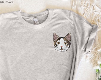 May include: Light gray t-shirt featuring an embroidered cat face on the chest. The cat has brown and white markings, with yellow eyes and a pink nose. The shirt has a crew neck and rolled-up sleeves. The brand name "MOD PAWS" is in the top left corner.