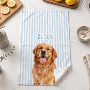May include: A rectangular kitchen towel featuring a light blue and white vertical striped pattern. The centre of the towel displays a realistic illustration of a golden retriever dog with its tongue out, above which the name BUDDY is printed.